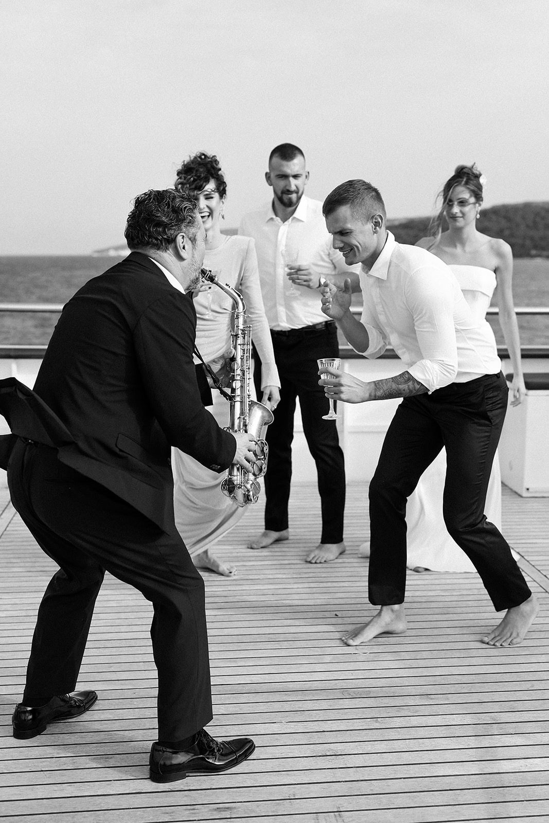 Party trenutak na palubi jahte – saksofonist i gosti plešu, more u pozadini, crno-bijela editorial fotografija